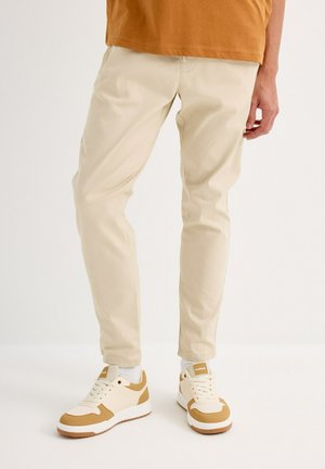 Pantalones color beige, ajustados y confeccionados en algodón, con bolsillos laterales. Combinados con zapatillas blancas y marrones que presentan un tejido texturizado y una suela de goma.