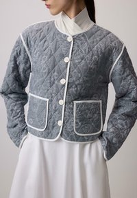 Pikovaná sivá bunda s bielou kvetinovou výšivkou, ktorá má zaoblený golier, predné gombíky a dve nášivkové vrecká s bielym lemom.