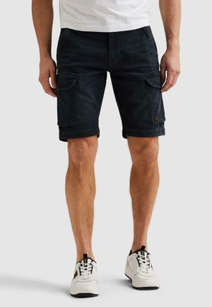 Mann trägt schwarze Cargo-Shorts mit Klappentaschen, weiße Sneakers und ein weißes Hemd vor einem schlichten hellen Hintergrund.