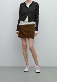 Schwarze Strickjacke mit Knöpfen, über einem weißen Oberteil getragen, kombiniert mit einem braunen Wildleder-Minirock und weißen Sneakern. Glatte Texturen und taillierte Passform.