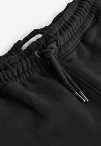 Schwarze Sweatpants mit strukturierter Taille, verstellbarem Kordelzug mit Metallspitzen und weichem Stoff. Keine sichtbaren Taschen oder Muster.