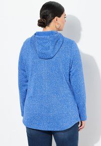 Blå huvtröja med en texturerad, fläckig design. Har en huva, långa ärmar och en rundad nederkant. Kombineras med mörka denimbyxor.