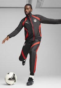 Survêtement noir avec des accents rouges, comprenant une veste zippée et un pantalon assorti, fabriqué en matériau léger. Joueur en train de dribbler un ballon de football.