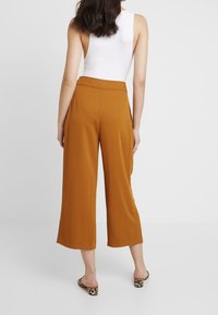 Kvinna med axel-långt hår som bär en vit ärmlös topp, rostfärgade vida cropped byxor och sandaler med klack i leopardmönster.