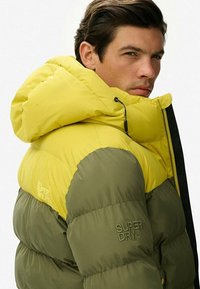 Kurtka puchowa z kolorowymi blokami, z żółtą kapturem i oliwkowym korpusem, z zamkiem z przodu i logo "SUPERDRY" z boku.