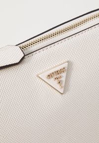 Weißes, strukturiertes synthetisches Handtasche mit goldfarbenen Beschlägen und einem dreieckigen Logo-Anhänger, auf dem "GUESS" in erhabenen Buchstaben steht. Mit Reißverschluss schließen.