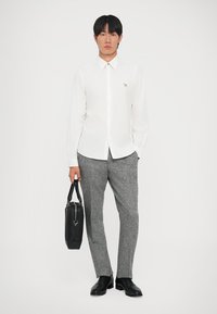 Camicia bianca con colletto, pantaloni grigi a trama, scarpe nere e una borsa nera. Modella in piedi contro uno sfondo semplice.