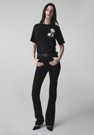 Frau trägt ein schwarzes T-Shirt mit weißem Blumenmuster, schwarzen Gürtel, schwarze taillierte Schlaghose und schwarze spitze Absätze, steht vor neutralem Hintergrund.