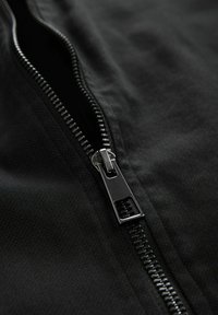 Schwarzer Stoff mit einem sichtbaren Metallreißverschluss. Der Reißverschluss verfügt über einen polierten Schieber und verzahnte Zähne, die sein elegantes Design hervorheben.