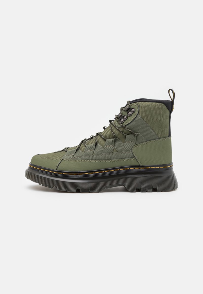 Botte montante vert olive avec semelle noire épaisse, coutures jaunes, dessus en tissu, lacets à l’avant et languette au dos.