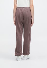 Pessoa a usar calças largas em tom malva com punhos elásticos e ténis brancos, vista de costas contra um fundo branco liso.