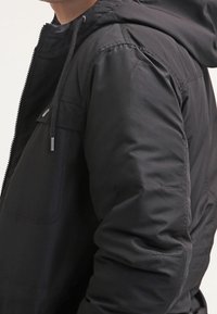 Svart huva jacka med dragkedja, quiltad textur, justerbara dragsnören och sidofickor. Tillverkad av ett hållbart, vattenavvisande material.