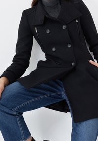 Cappotto peacoat nero con bottoni doppi, tasche con zip e un design in tessuto testurizzato. Abbinato a jeans blu, che mostrano una vestibilità su misura.