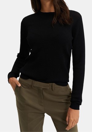 Femme portant un pull à manches longues noir texturé et un pantalon vert olive ceinturé, une main dans la poche, debout devant un fond uni.