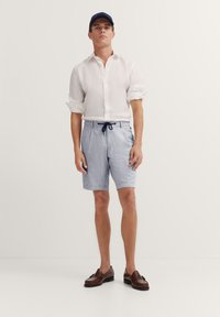 Leichtes weißes Leinenhemd, blau gestreifte Shorts mit Kordelzug, braune Lederloafers und eine marineblaue Mütze. Neutraler Hintergrund.