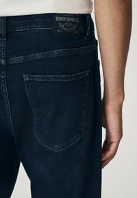 Jeans din denim albastru închis cu un buzunar din spate vizibil și un model cusut. Banda de talie are o etichetă neagră pe care scrie "DENIM SUPPLY Co."