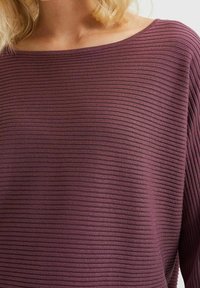 Nainen, joka on pukeutunut ribbittyyn, pitkähiharaiseen, mauvenväriseen venepääntie-paitaan, ja hänen vaaleat hiuksensa näkyvät osittain kaula-aukosta.