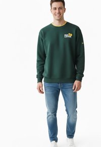 Joven sonriente, vestido con una sudadera verde oscuro con el texto "SEA LOVER", jeans azules y zapatillas blancas sobre un fondo blanco.