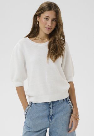 Jeune femme aux longs cheveux bruns portant un pull blanc à manches bouffantes, un jean bleu avec des poches ornées, des boucles d’oreilles créoles dorées et des bracelets.