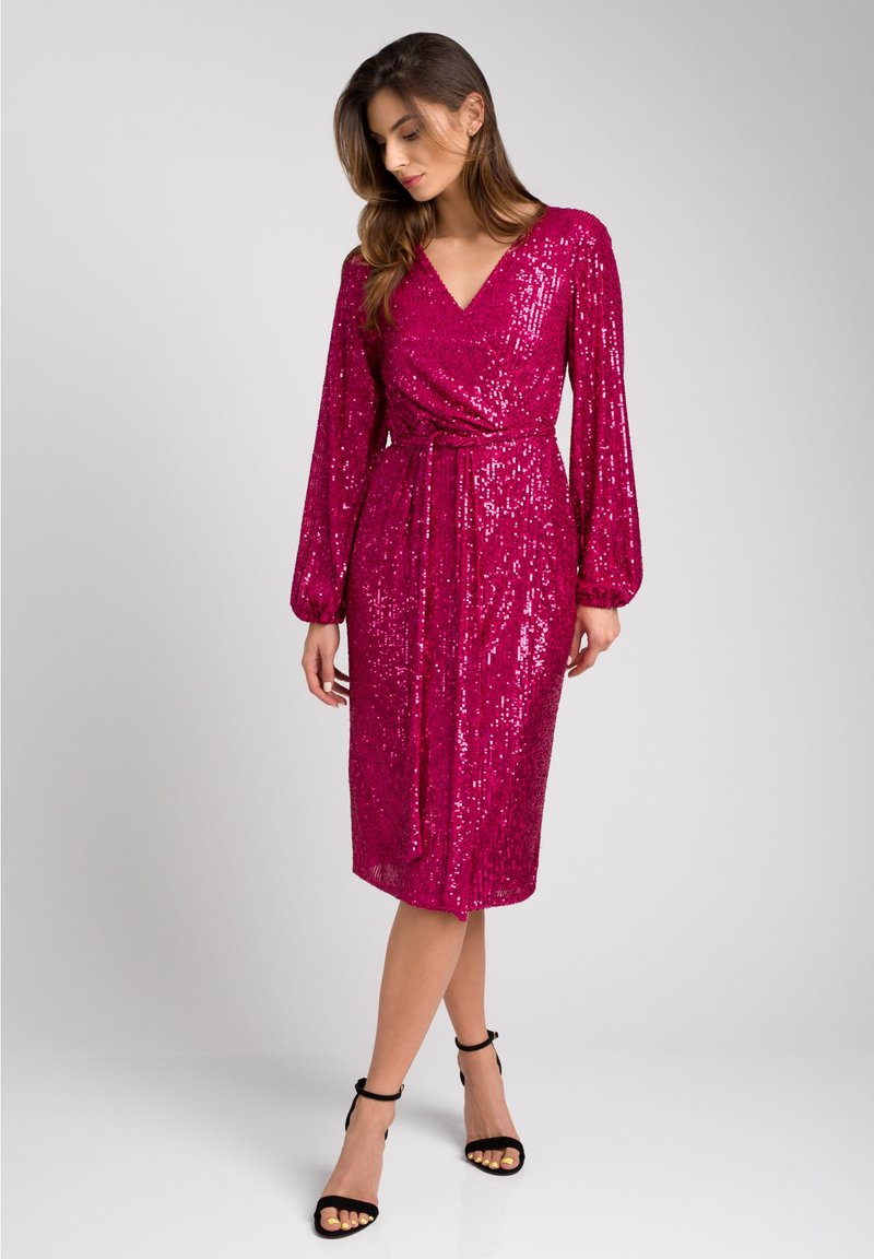 Fuksjowa sukienka z cekinami, z długimi rękawami, fasonem kopertowym, dekoltem w serek i materiałowym paskiem; długość do kolan z gładką teksturą. Czarne sandały na ramiączkach.