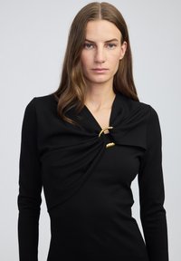 Zwarte jurk met een gedrapeerd voorpand, lange mouwen en een gouden accentdetail bij de halslijn, die de op maat gemaakte pasvorm benadrukt.