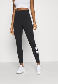 Svarta leggings i stretchmaterial med hög midja och en vit Nike-logotyp på vänster ben. Bärs tillsammans med en vit crop top och sneakers.