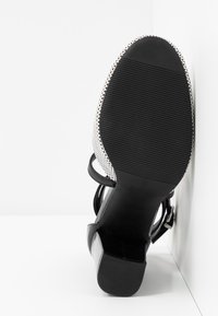 Chaussure à talon noir avec une semelle texturée, une finition brillante en vernis, et un dessus à motifs avec des sangles ajustables. Vue de côté sur fond blanc.