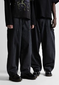 Juodos džinsinės cargo kelnės turi laisvą siluetą, matomas kišenes ir siuvinėjimą. Renkamos su juodais batais, papildytos grafika puoštomis juodomis marškinėliais.