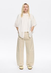 Camicia larga bianca a righe con bottoni, top color crema, pantaloni beige a gamba larga e scarpe giallo chiaro con lacci e dettaglio della suola a contrasto.