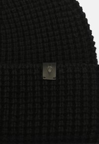 Black knitted beanie with a ribbed texture, featuring a small black metal label with a skull design on the folded hem.