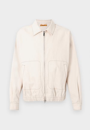 Veste beige zippée avec un col, deux grandes poches avant, un ourlet élastiqué et des détails de couture contrastants marron.