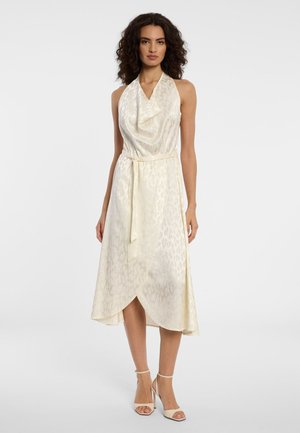 Frau mit lockigem Haar, die ein ärmelloses, elfenbeinfarbenes Satinkleid in Midilänge mit drapiertem Ausschnitt, seitlichem Schlitz, Taillengürtel und beigen High-Heel-Sandalen trägt.