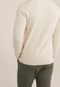 Light beige textured sweater with a honeycomb pattern, ribbed hem, and cuff details, paired with green trousers. Back view.