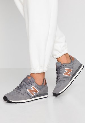 Person iført hvide joggingbukser og grå New Balance-sneakers med roseguldaccenter, vist fra anklen nedad på en lys gulv.
