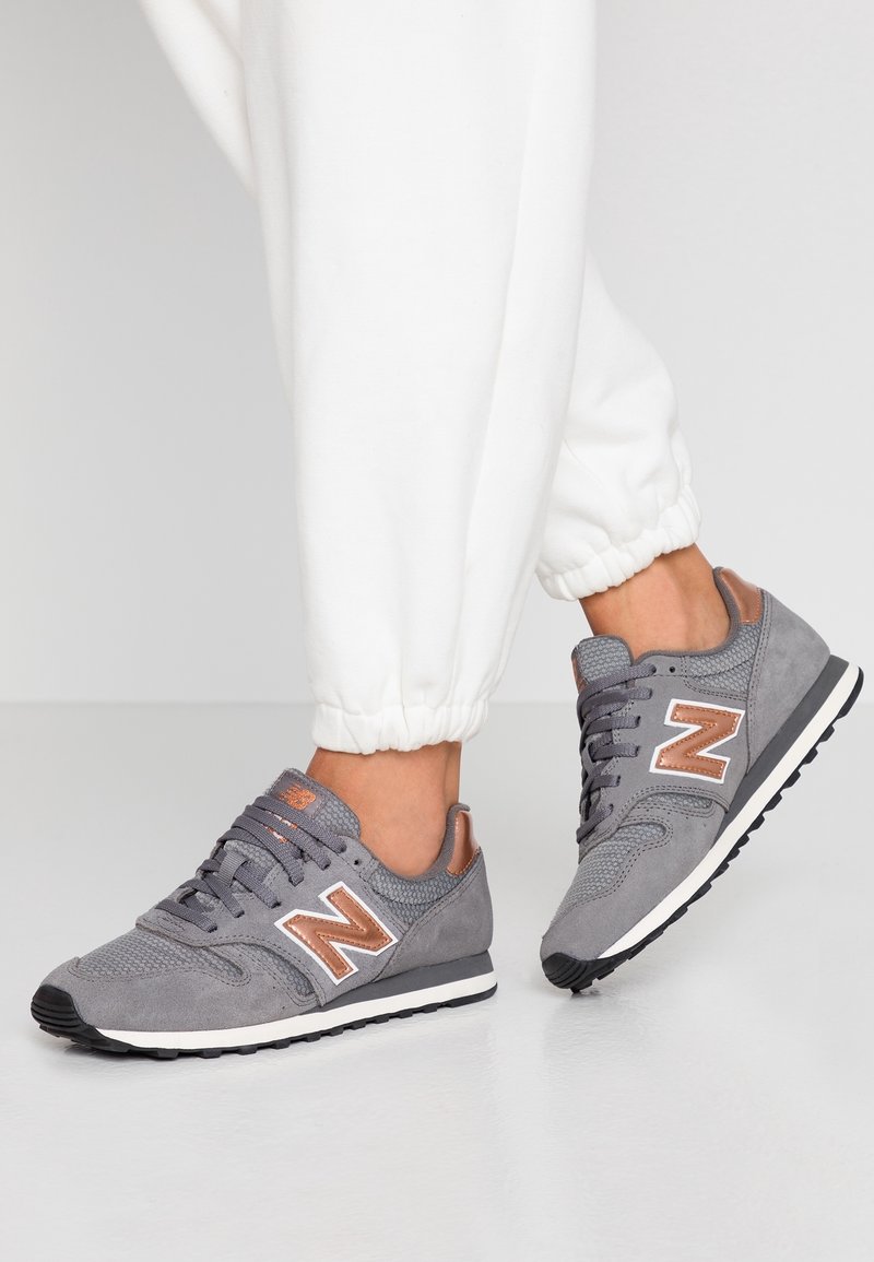 Personne portant un pantalon de jogging blanc et des baskets New Balance grises avec des accents en or rose, montrée de la cheville vers le bas sur un sol clair.