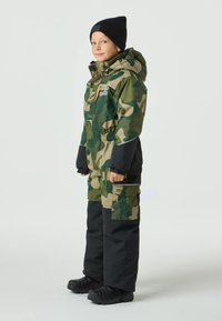 Kombinezon zimowy w kamuflażu z kapturem, elastyczną talią i czarnymi akcentami. Posiada kieszenie na zamek i wodoodporne materiały. Czarne buty.