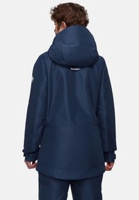 Marineblaue wasserdichte Jacke mit einer großen, verstellbaren Kapuze, elastischen Bündchen und einem Logo auf dem oberen Rücken. Glatte Textur, knielanges Design.