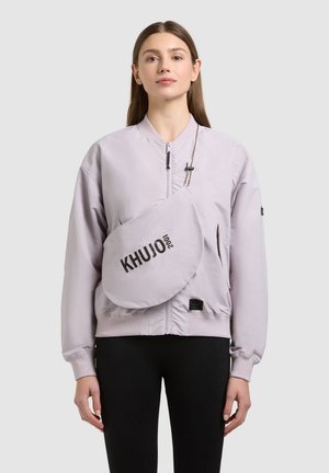 Žena v svetlofialovej bomber bunde a s kabelkou cez rameno s nápisom "KHUJO 2001", stojaca pred jednoduchým pozadím.