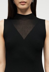 Femme portant un haut noir sans manches côtelé avec un panneau transparent en forme de V au niveau de la poitrine et un col montant.