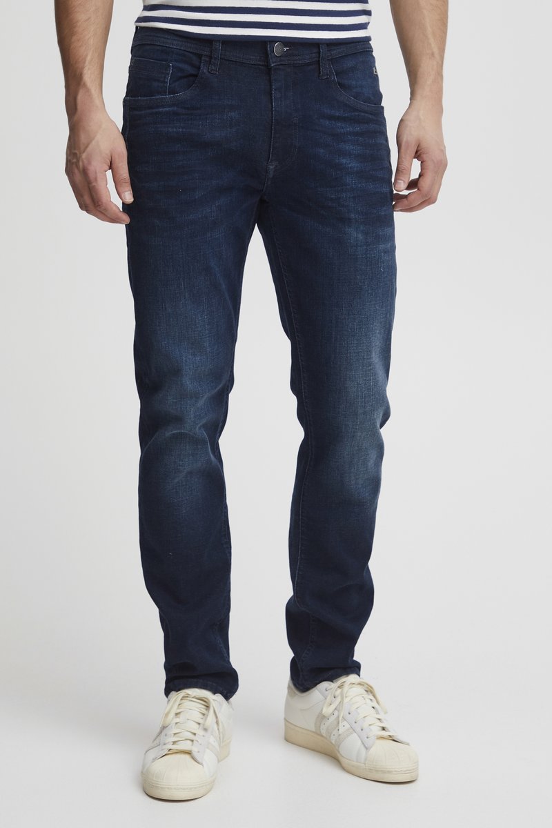 Muž v tmavomodrých slim fit džínsoch a bielych teniskách stojí pred jednoduchým svetlým pozadím.