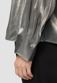 Metalen zilveren shirt met een gladde textuur, lange mouwen met geraffineerde details en knoopmanchetten. Zwarte stof gedeeltelijk zichtbaar.