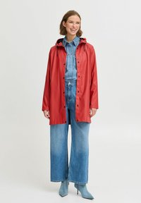 Impermeabil roșu cu glugă, cu nasturi tip snap, croială supradimensionată, purtat peste o ținută din denim, cu blugi evazați și cizme anklet albastre.