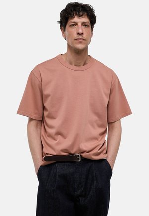 Homme aux cheveux bouclés portant une chemise ample à manches courtes brun pâle rentrée dans un pantalon sombre avec une ceinture noire, debout les mains dans les poches.