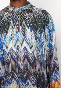 Mönstrad blus i siden med flerfärgade zigzag-ränder, med hög halsringning och liten öppning samt plisserad design, främst i blått, grönt och svart.