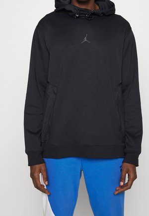 Mann trägt einen schwarzen Hoodie mit grauem Jumpman-Logo und blaue Hose mit weißen und schwarzen Details, steht vor schlichtem Hintergrund.