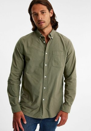 Olivenfarvet skjorte med knapper og en enkelt brystlomme, justerbare manchetter og et blødt, struktureret stof. Kombineret med blå jeans.