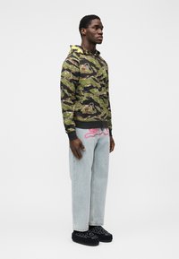 Φούτερ με κουκούλα που διαθέτει μοτίβο παραλλαγής σε πράσινο, καφέ και μαύρο χρώμα. Συνδυάζεται με ανοιχτόχρωμο μπλε τζιν και μαύρα παπούτσια τύπου slip-on με σχέδια.