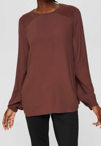 Blusa marrone a maniche lunghe con design plissettato sulle spalle, scollo rotondo, tessuto morbido e polsini aderenti, abbinata a pantaloni neri.