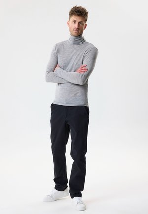 Vyras stovintis su sukryžiuotomis rankomis, dėvintis šviesiai pilką golfą, juodus kelnes ir baltus sportinius batelius ant vienspalvio baltos fono.