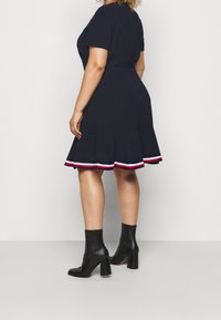 Persona con curvas vestida con un vestido de manga corta en color marino con un dobladillo acampanado y detalles a rayas, combinando con botas negras hasta el tobillo, de pie frente a un fondo neutro.